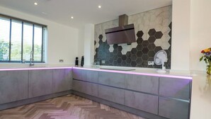 A modern kitchen with a herringbone wood floor, a built-in grey island with a pink LED under-cabinet lighting, a stainless steel range hood, and a hexagonal tile backsplash. - Carnon Gwel, Devoran (Truro)