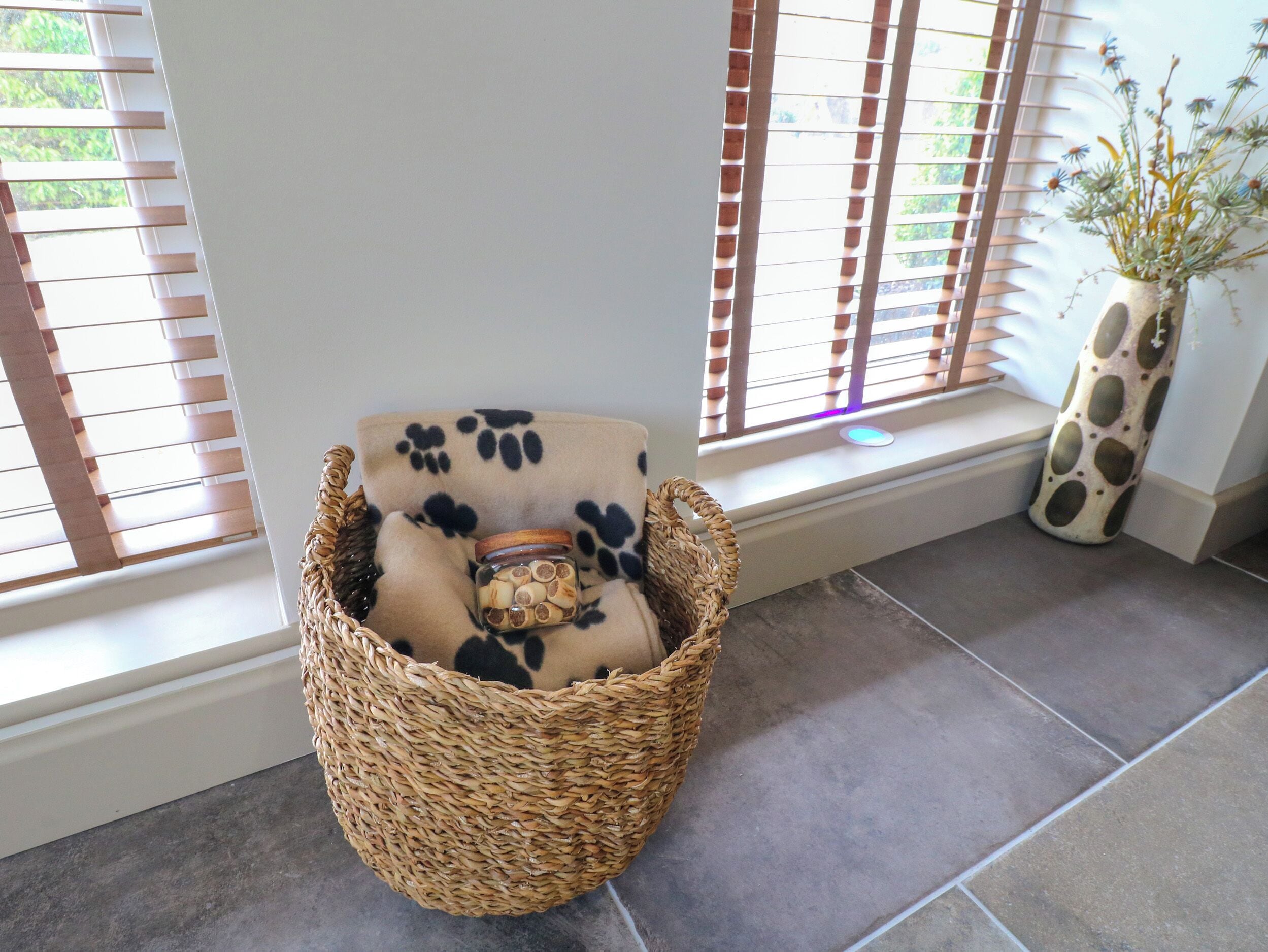 A wicker chair with a cushion and a small jar of snacks, positioned near a window with blinds.