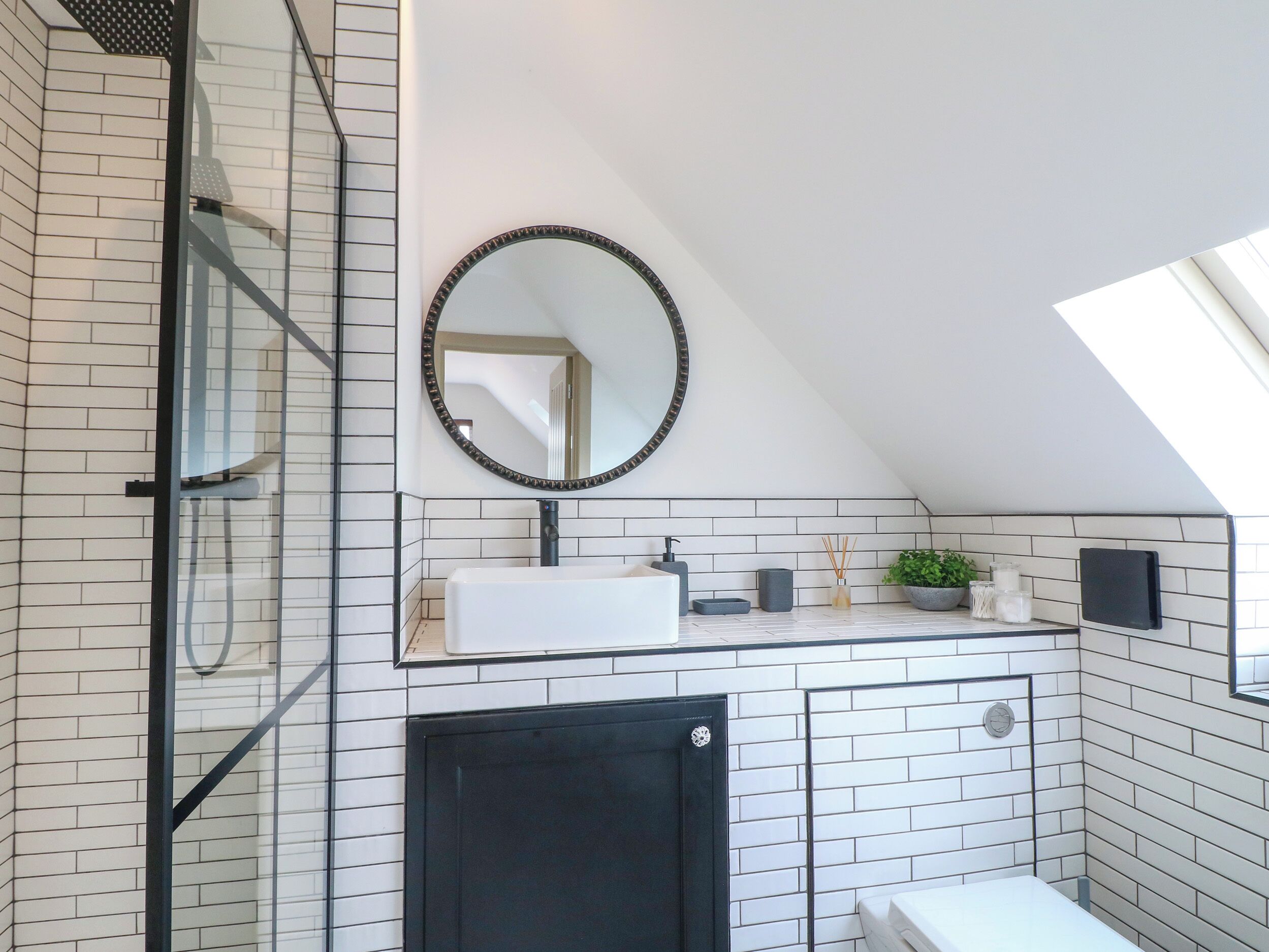 Un baño moderno con tragaluz, azulejos blancos tipo metro, un mueble negro y un espejo redondo.