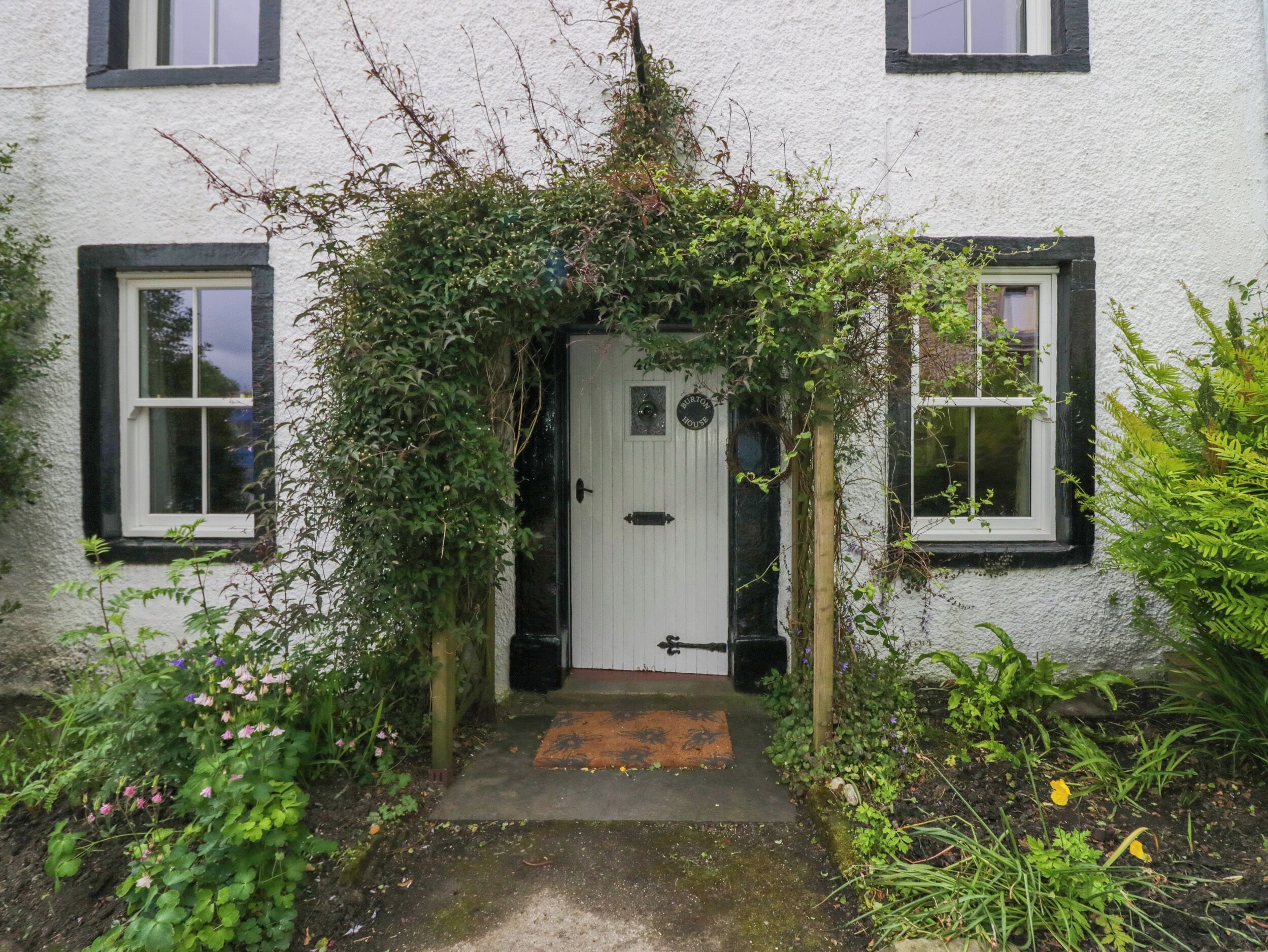 A white house with a white door and black-framed windows, surrounded by greenery and a small garden.