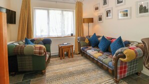 Ein gemütliches Wohnzimmer mit Sofa, Sessel und einem kleinen Tisch. Vorhänge, eine Lampe und gerahmte Bilder schmücken die Wand.