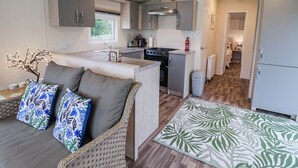 A compact kitchen with a stove, sink, and cabinets. There is a small dining area with a sofa and cushions. - The Hoot (Stowmarket)