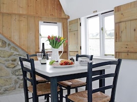 Una mesa de comedor con mantel blanco, rodeada por cuatro sillas de madera oscura con asientos de mimbre. En la mesa hay una botella de vino, una jarra blanca con flores de colores, y un plato con uvas y tomates.