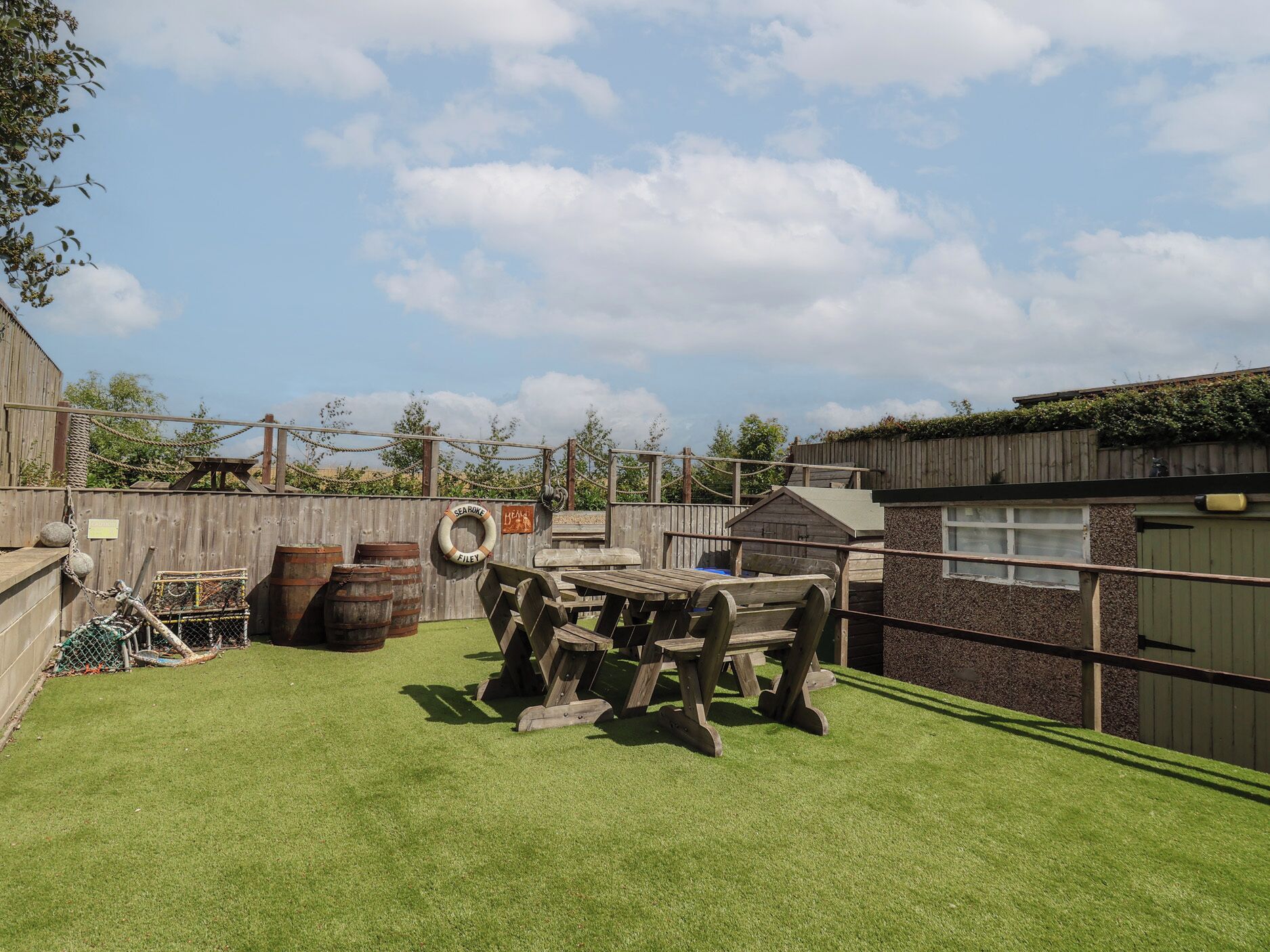 A fenced outdoor area with a wooden table and chairs, barrels, and a rope swing.