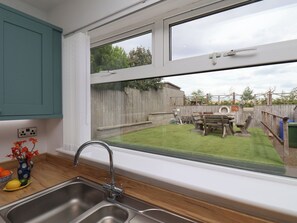 A kitchen with a large window, a sink, and a view of a backyard with a wooden fence and a barbecue area. - Sea Roke (Filey)
