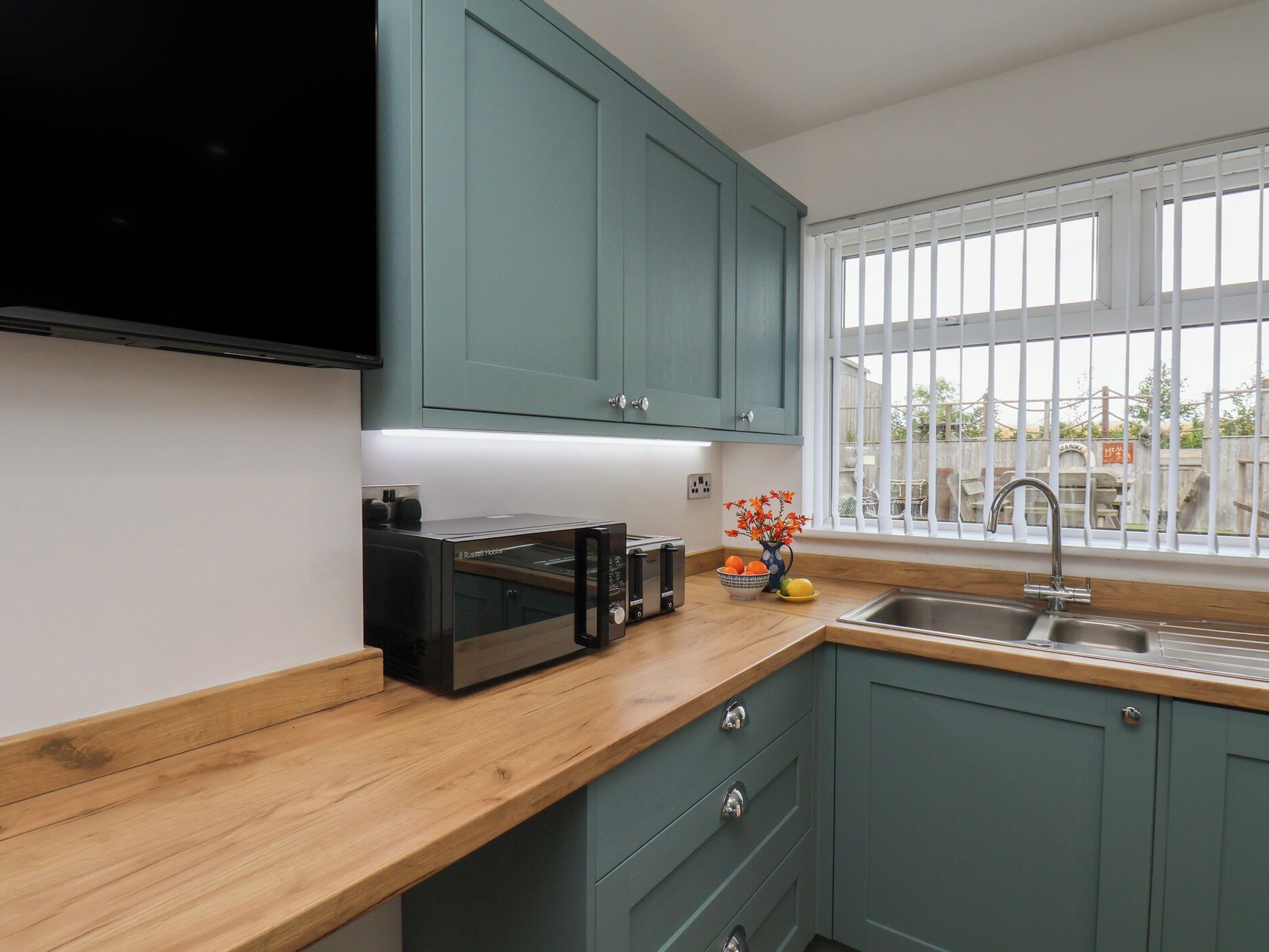 A kitchen with teal cabinets, a wooden countertop, a built-in microwave, a toaster, and a sink.
