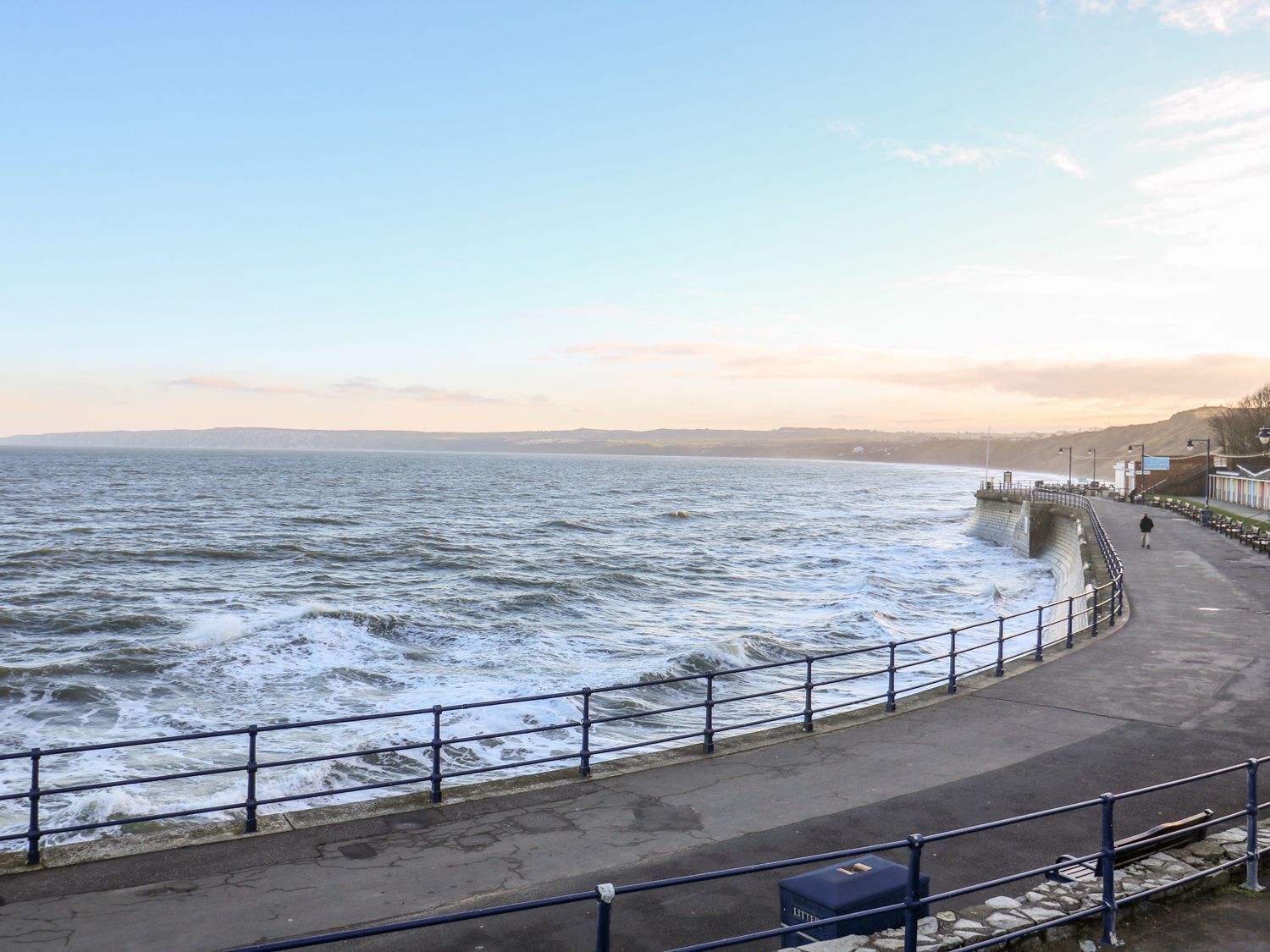 A coastal promenade with a stone sea wall, a paved walkway, and a clear view of the sea.
