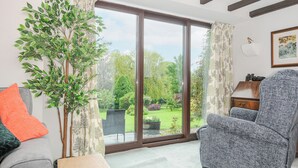 A living room with a large sliding glass door leading to a garden, a grey sofa with an orange pillow, a grey armchair, and a small wooden table with a laptop. - Manor Barn (Saltash)
