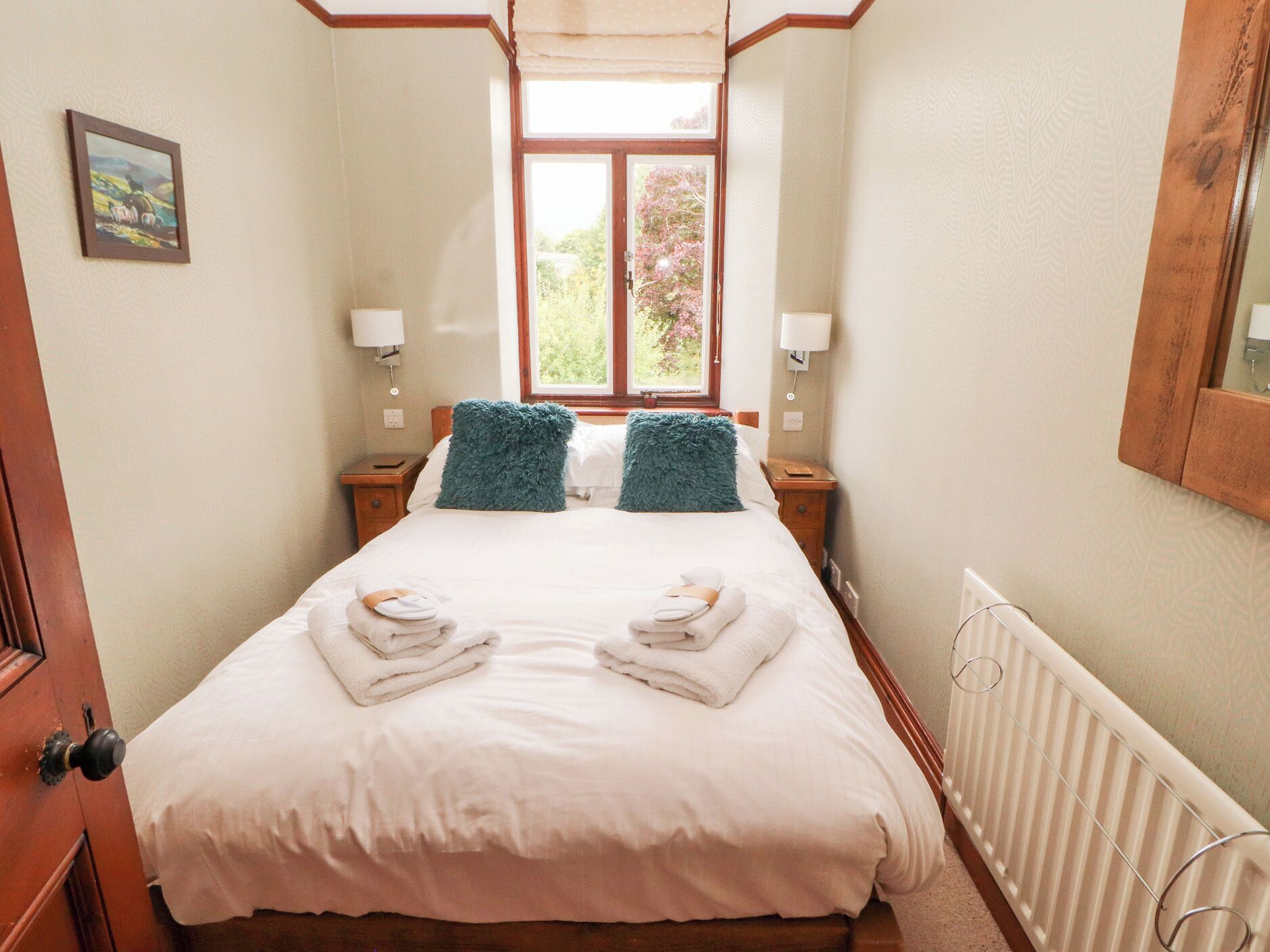 A narrow hotel room with a large bed, two nightstands, a window with a view of greenery, and a radiator.