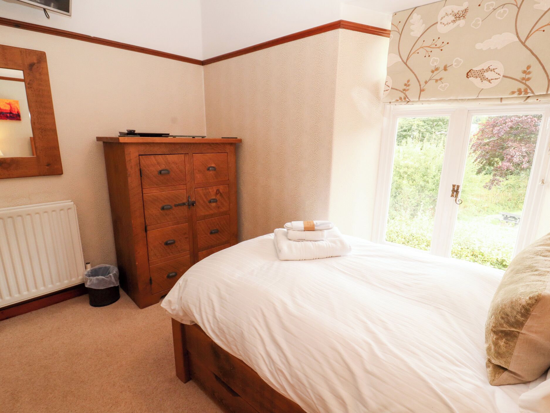 A bedroom with a wooden dresser, a radiator, a window with a view of greenery, and a neatly made bed.