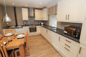 A modern kitchen with white cabinets, a wooden dining table set for four, and a built-in oven. - Tremornah (Penzance)