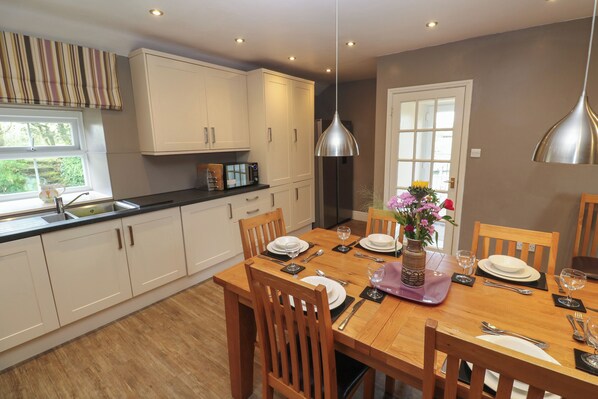 A modern kitchen with white cabinets, a wooden dining table set for four, and a large window providing natural light. - Tremornah (Penzance)