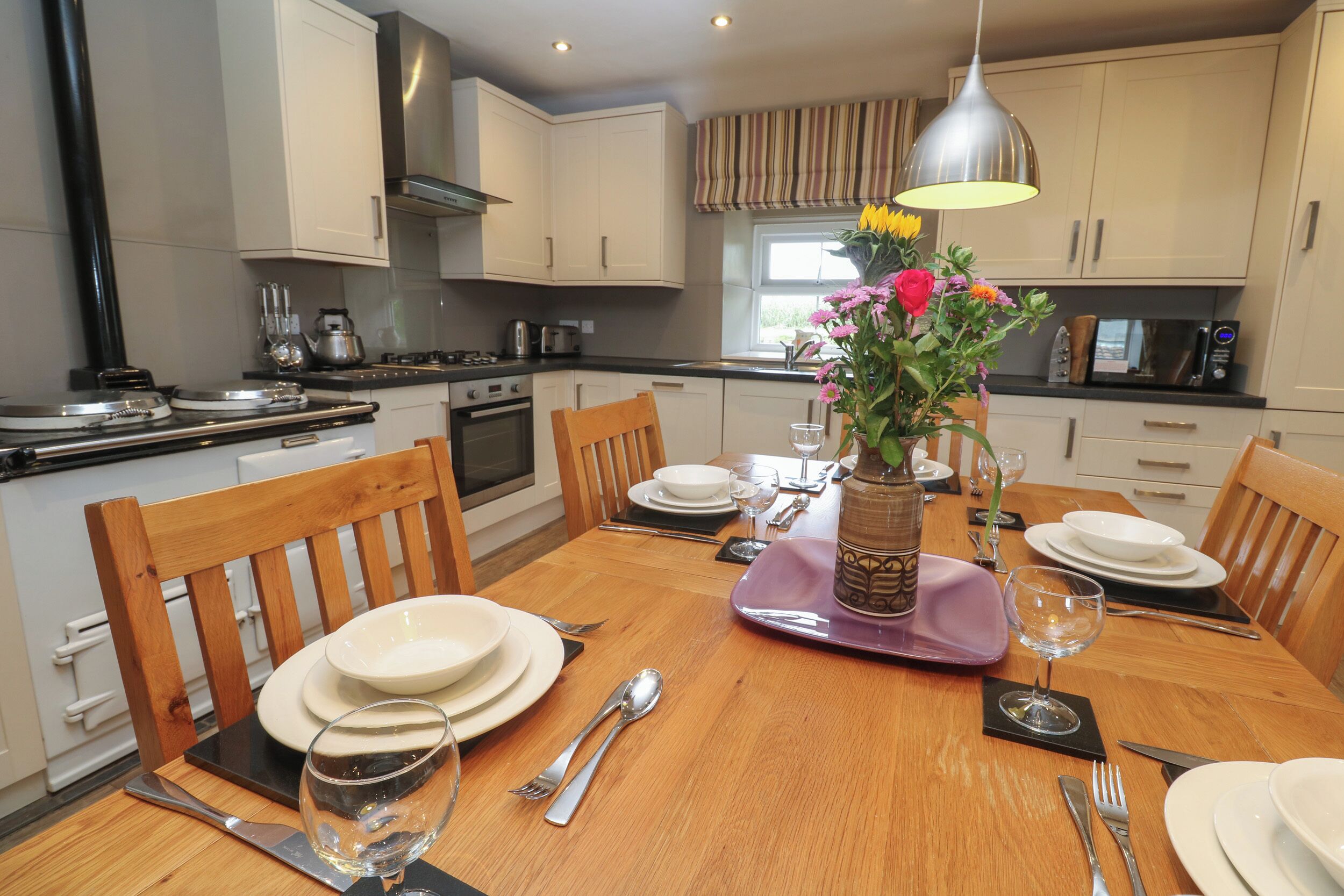 A dining table set with plates, cutlery, and a vase of flowers in a modern kitchen with white cabinets and a window.