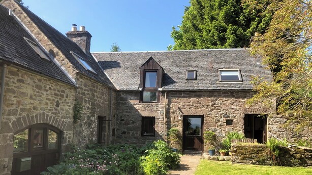 Ein Steinhaus mit mittlerem Eingang, einer kleinen Veranda und einem Garten mit Kübelpflanzen.