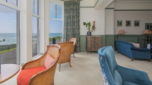 A room with large windows, wicker chairs, a blue armchair, and a view of the sea. - Daphne (Helston)