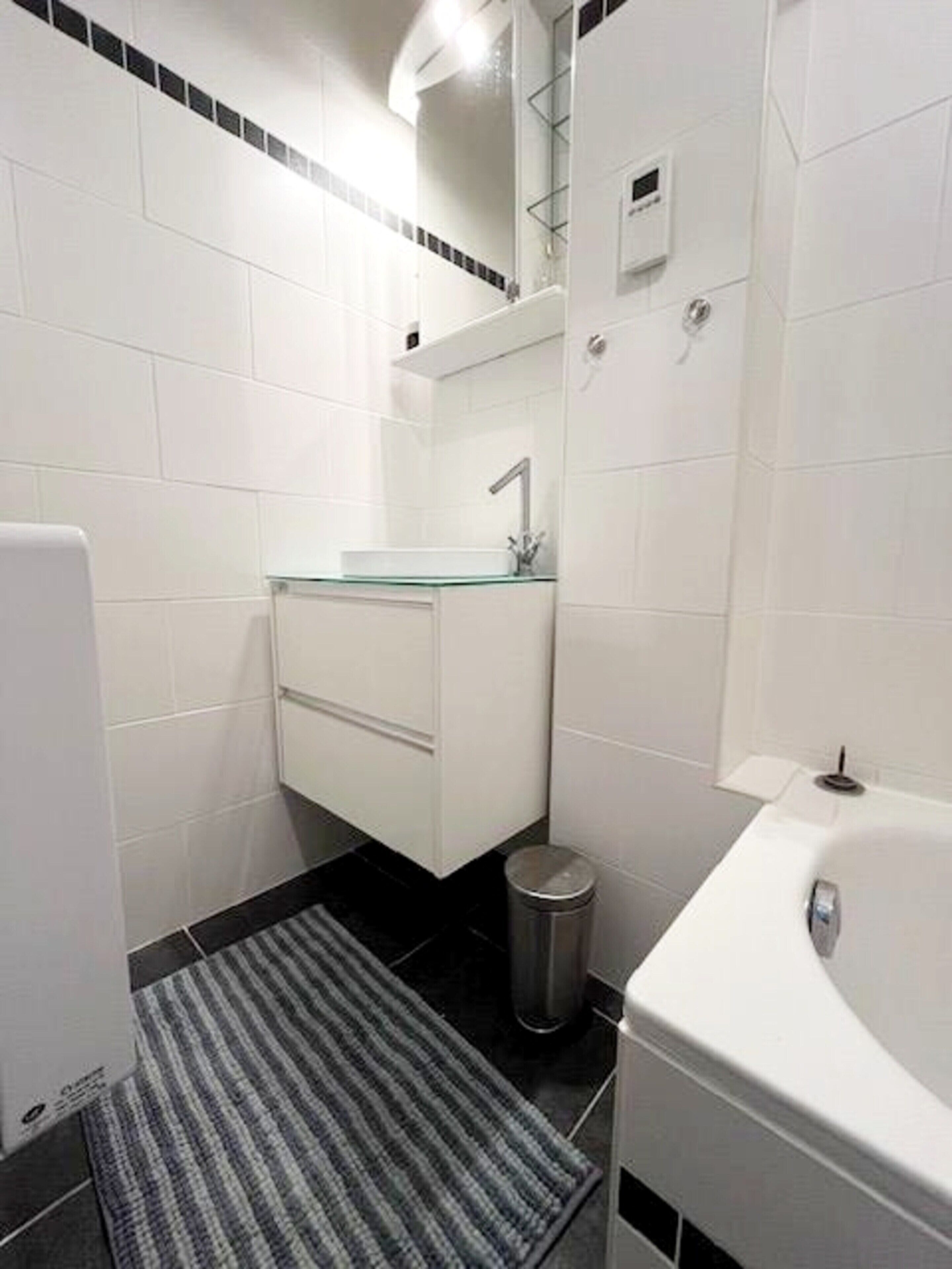 Ein Badezimmer mit einem Glaswaschbecken, weißen Schränken, einer schwarzen Fußmatte und einer weißen Badewanne.
