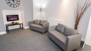 A modern living room with two grey sofas, a black clock, a flat-screen TV, and a radiator. - Apartment 1 (Llandudno)