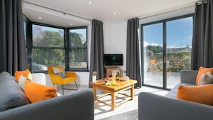 A modern living room with two grey sofas, a yellow chair, a wooden coffee table with snacks, and a TV. Large windows offer a view of the outdoors. - Nova Cottage (St Ives)