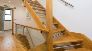 A modern wooden staircase with glass railings and metal handrails, leading to an upper floor. - Nova Cottage (St Ives)