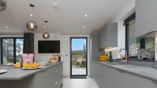 A modern kitchen with grey cabinets, a flat-screen TV, and a dining area with a view. - Nova Cottage (St Ives)