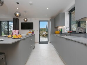 A modern kitchen with grey cabinets, a flat-screen TV, and a dining area with a view. - Nova Cottage (St Ives)