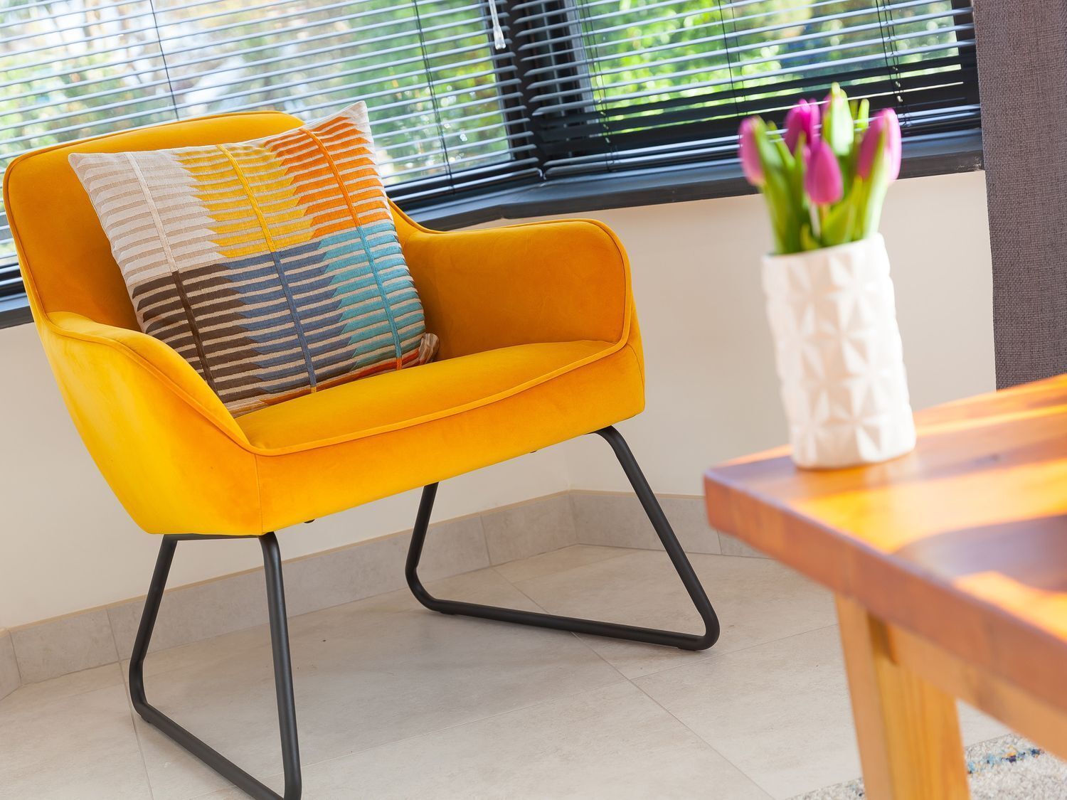 A bright yellow chair with black metal legs and two patterned cushions, positioned near a window with blinds and a vase of flowers on a wooden table.
