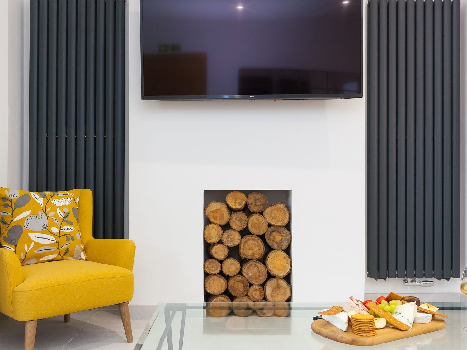 Un salón moderno con un sillón amarillo, una mesa de centro de vidrio, una exhibición de troncos de madera y una tabla de quesos.