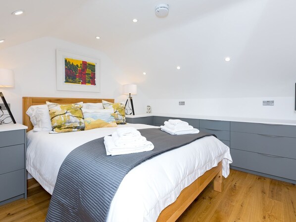 A modern hotel room with a large bed, grey and white bedding, a colorful painting on the wall, and a wooden floor.