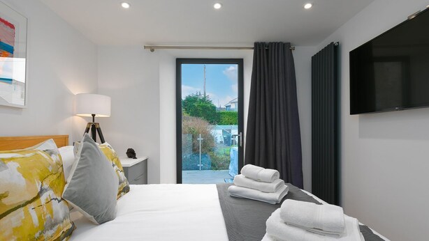 Moderni hotellihuone, jossa on sänky, yöpöytä lampun kanssa, televisio ja näkymä puutarhaan lasiovien kautta.