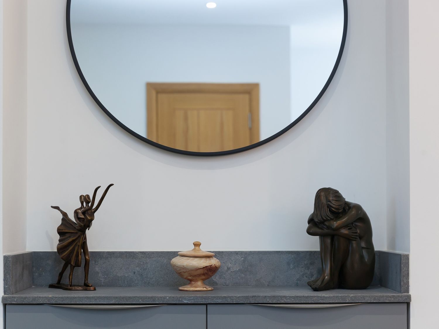 Een badkamerkastje met een spiegel, een klein beeld van een figuur met opgetrokken armen, een decoratieve vaas en een zittend figuur.