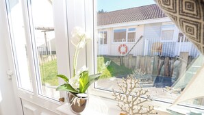 A window with a white orchid in a silver pot and a small tree sculpture on a white shelf.