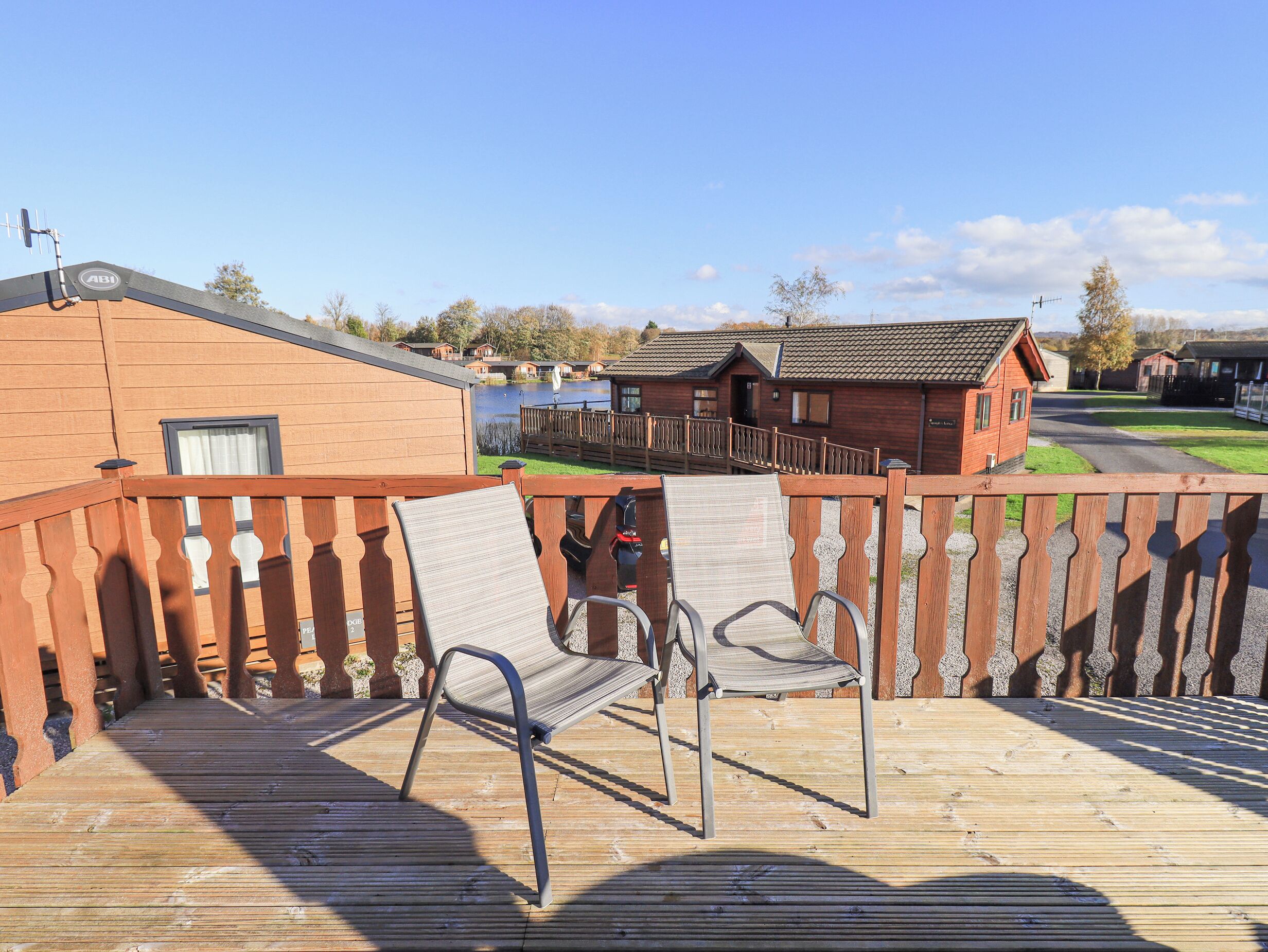 Eine Holzterrasse mit zwei Stühlen, einem Holzzaun und Blick auf einen See sowie Blockhäuser im Hintergrund.