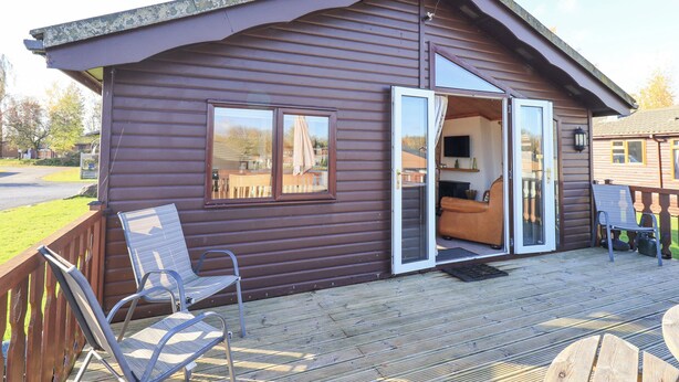 A wooden cabin with a deck, a sliding glass door leading to a furnished interior, and a small outdoor seating area. - Kellet Lodge (Carnforth)
