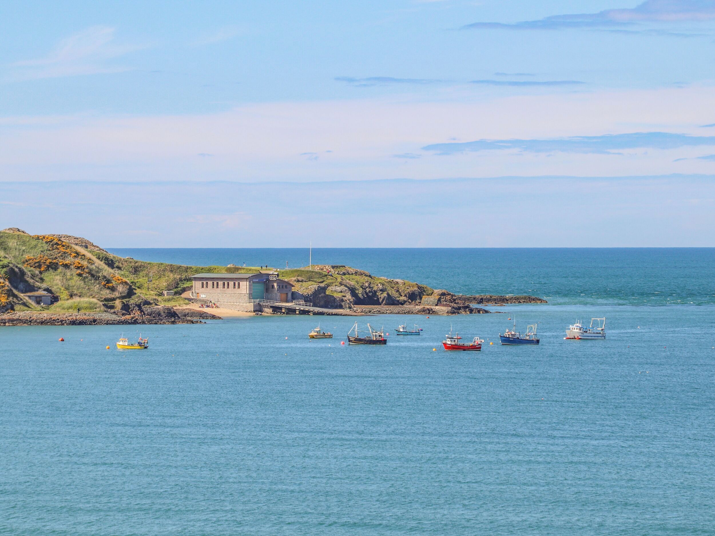 Un paysage côtier avec un petit port, plusieurs bateaux et un phare situé sur un promontoire rocheux.