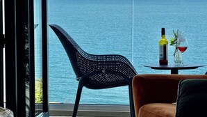 Une chambre moderne avec une grande fenêtre donnant sur la mer, une chaise noire perforée, une table ronde avec une bouteille et un verre, et un canapé marron.
