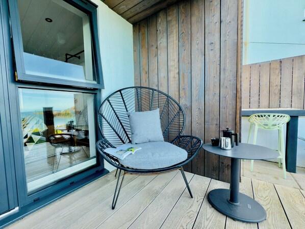 A modern outdoor seating area with a wicker chair and a small round table, wooden deck flooring, and a view through a large window.