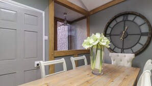 Un comedor con mesa de madera, sillas blancas, un reloj de pared grande y un florero con flores.