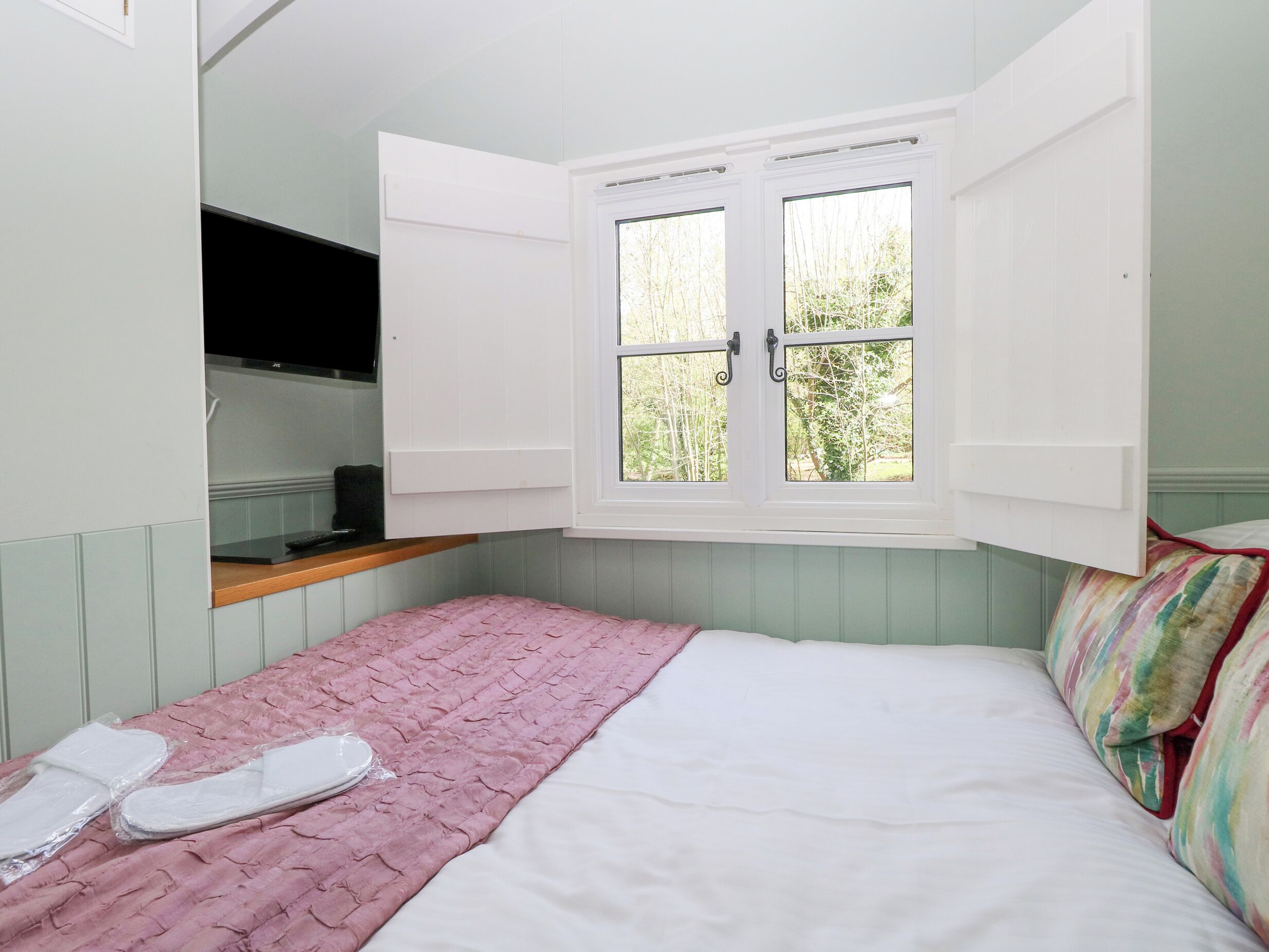Una camera da letto con un letto, una coperta rosa, una finestra con persiane aperte e un angolo cottura.
