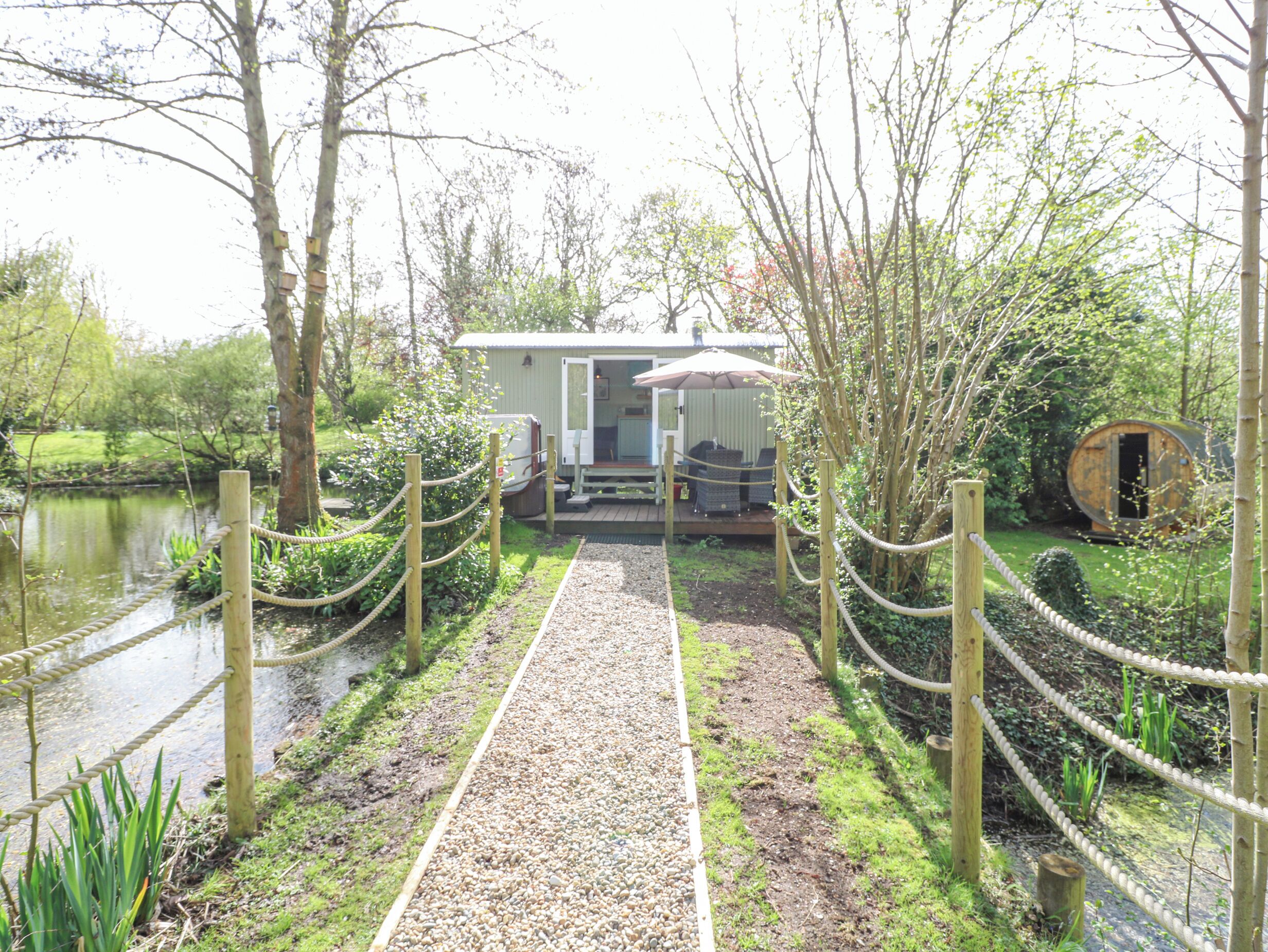 Un sendero conduce a una cabaña rústica con terraza y mobiliario exterior, rodeada de árboles y un pequeño estanque.