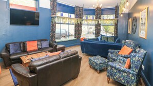 A living room with blue walls, a large window with curtains, a brown leather sofa, a blue patterned armchair, and a wooden coffee table. - Blue Ron's (High Peak)