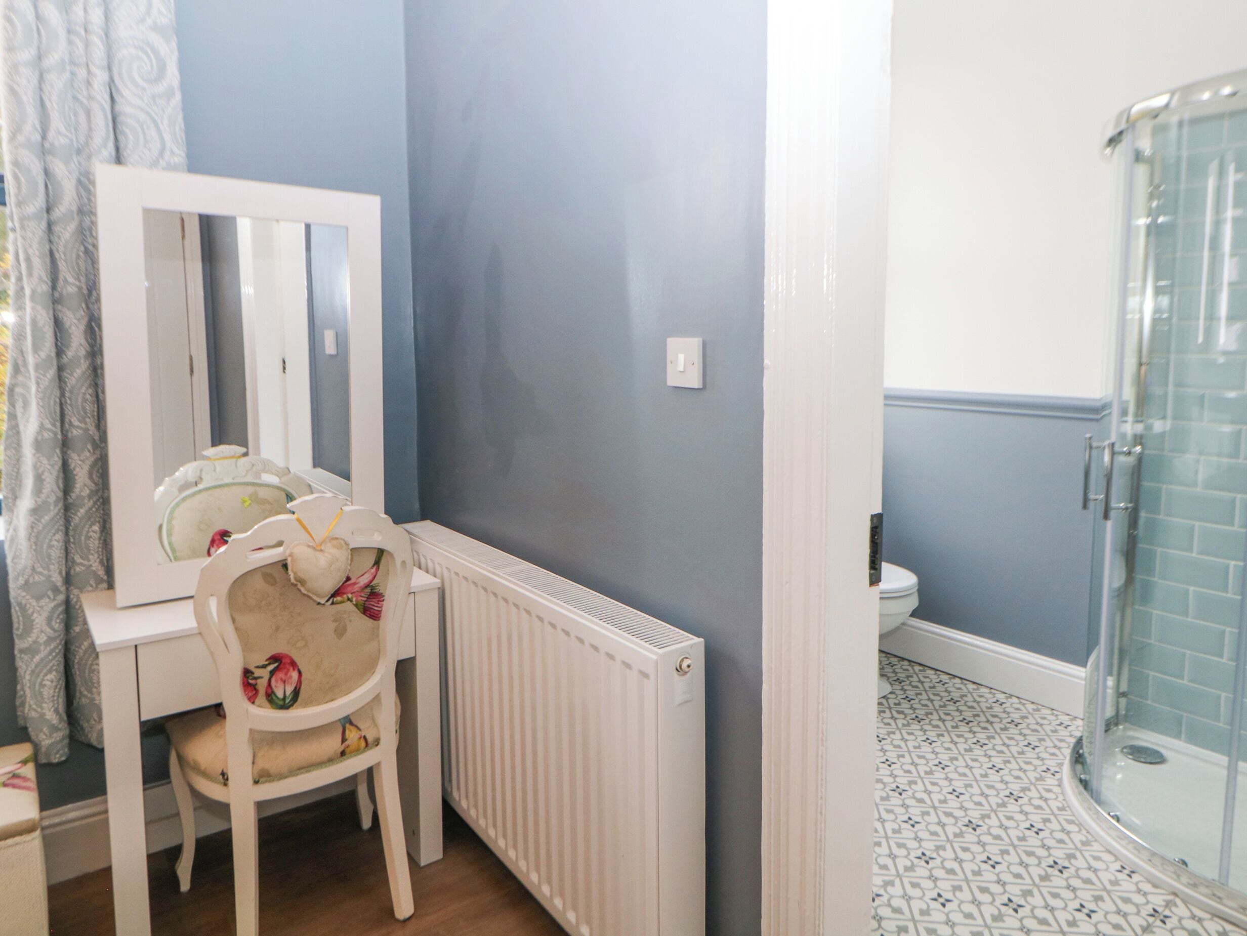 A bathroom with a white radiator, a mirror, a chair with floral upholstery, and a shower area with blue-tiled walls.