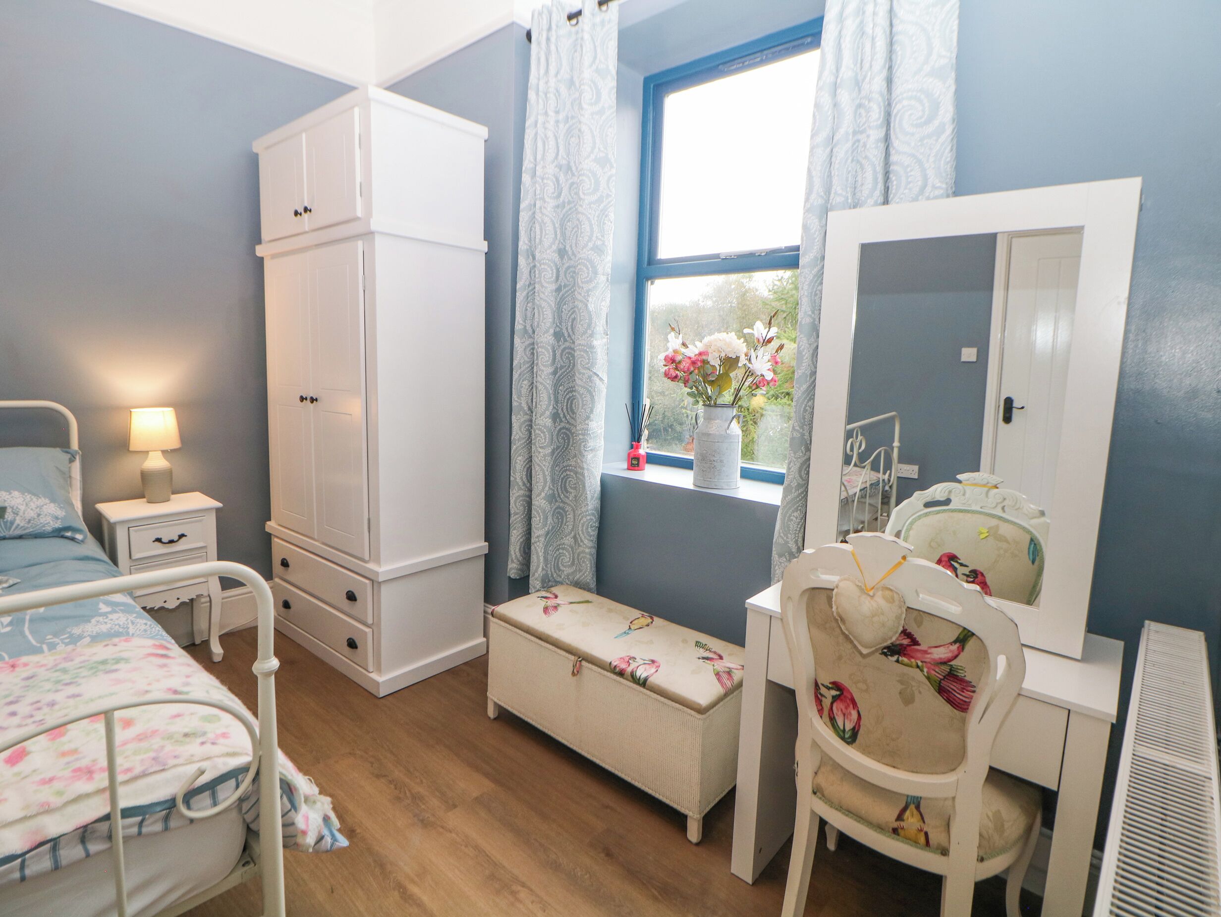 A bedroom with a bed, a nightstand, a wardrobe, a desk with a mirror, and a window with curtains.