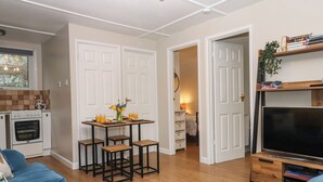 A living room with a blue sofa, a wooden dining table with four chairs, a TV stand with a flat-screen TV, and a kitchen area with an oven and a window. - 2 The Glade (Bude)