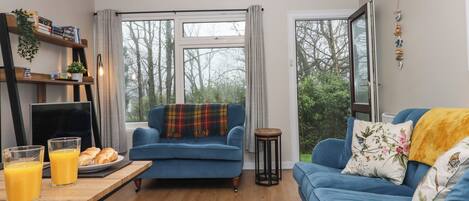 A modern living room with a blue sofa, a dining table with breakfast items, and large windows offering a view of trees.