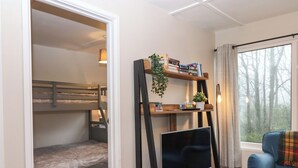 A room with a bunk bed, a TV on a wooden stand, a bookshelf with books and a plant, and a window with a view of trees. - 2 The Glade (Bude)