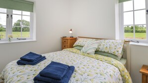A bedroom with a bed, two windows with green curtains, a bedside table with a lamp, and a view of a green landscape. - Sunset Cottage (Towcester)