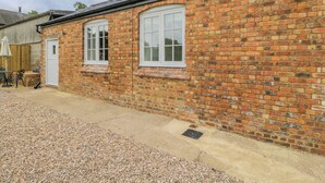 A brick building with two windows and a door, a gravel driveway, and a wooden fence in the background. - Sunset Cottage (Towcester)