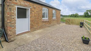A brick building with a white door and windows, a gravel driveway, and a wooden fence. - Sunset Cottage (Towcester)