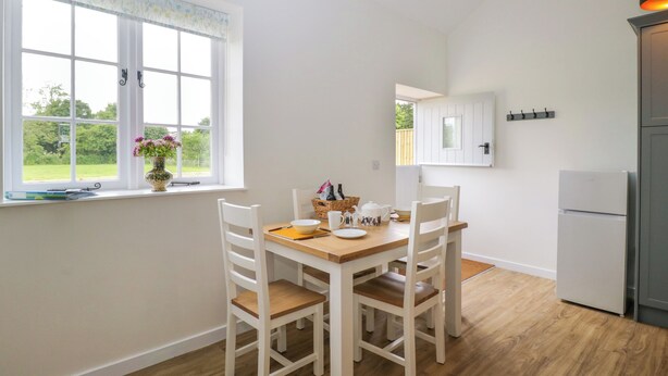 A kitchen with a dining table, chairs, a window with a view of greenery, a white cabinet, and a refrigerator. - Sunset Cottage (Towcester)