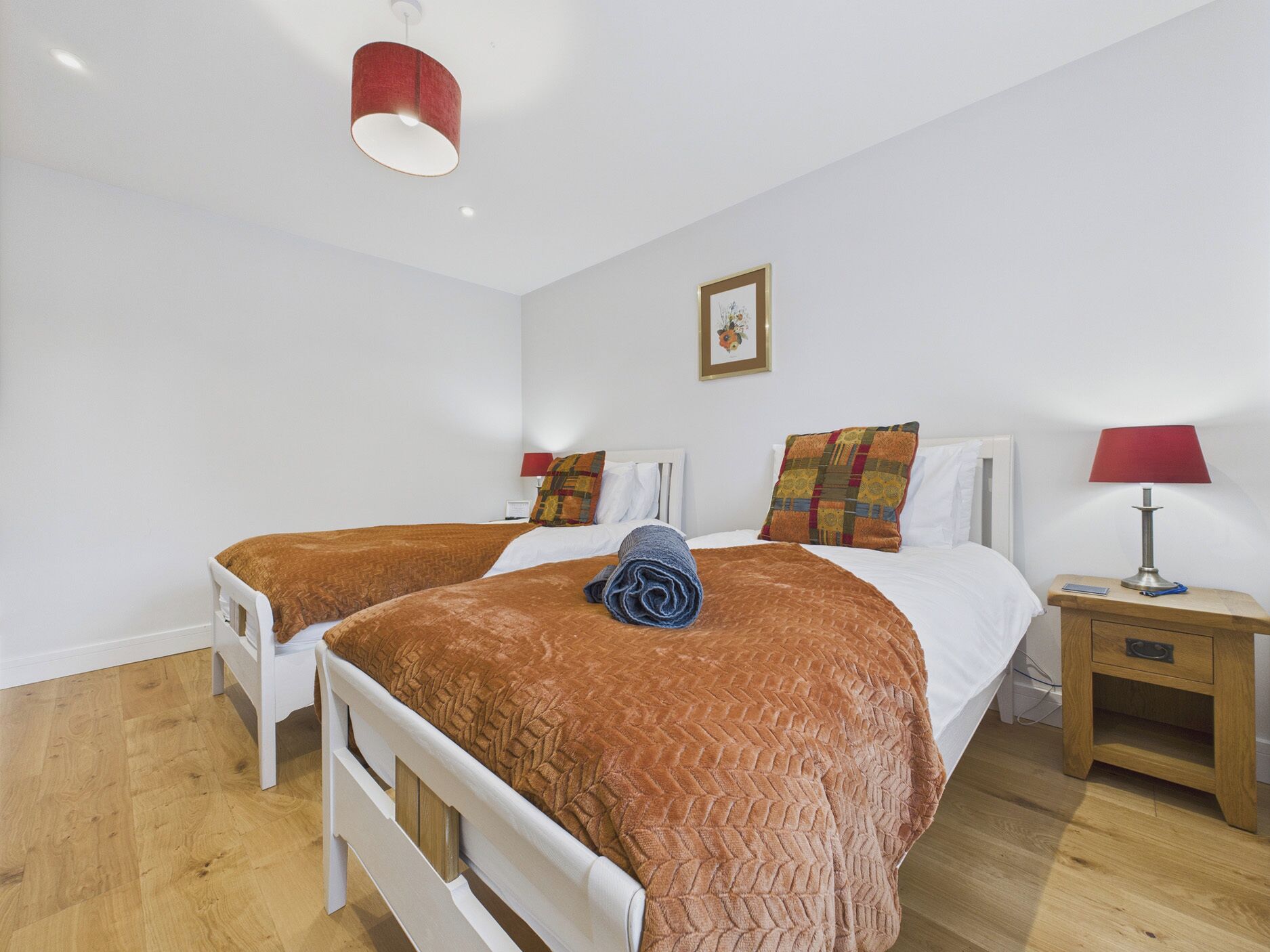 A hotel room with two beds, a wooden floor, a red lamp on the nightstand, and a framed picture on the wall.